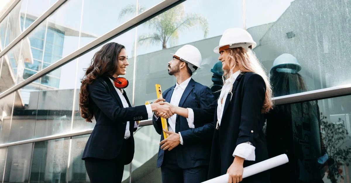 Three people, two wearing hard hats, shaking hands in front of a glass building