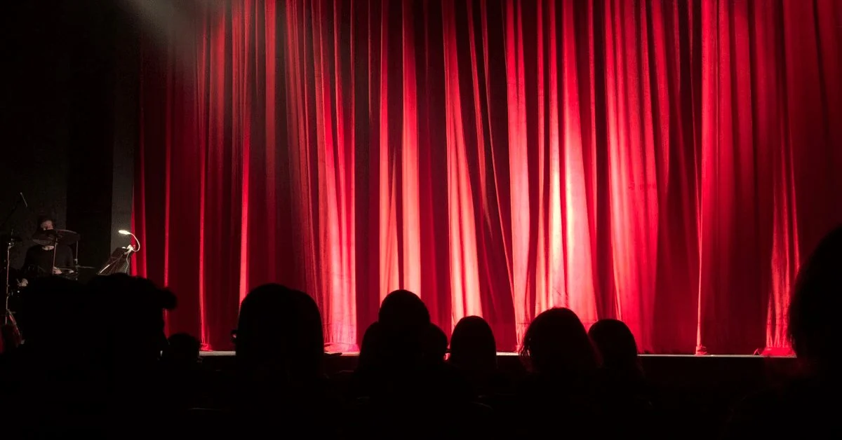 Red stage curtain under spotlights, audience silhouettes in the foreground